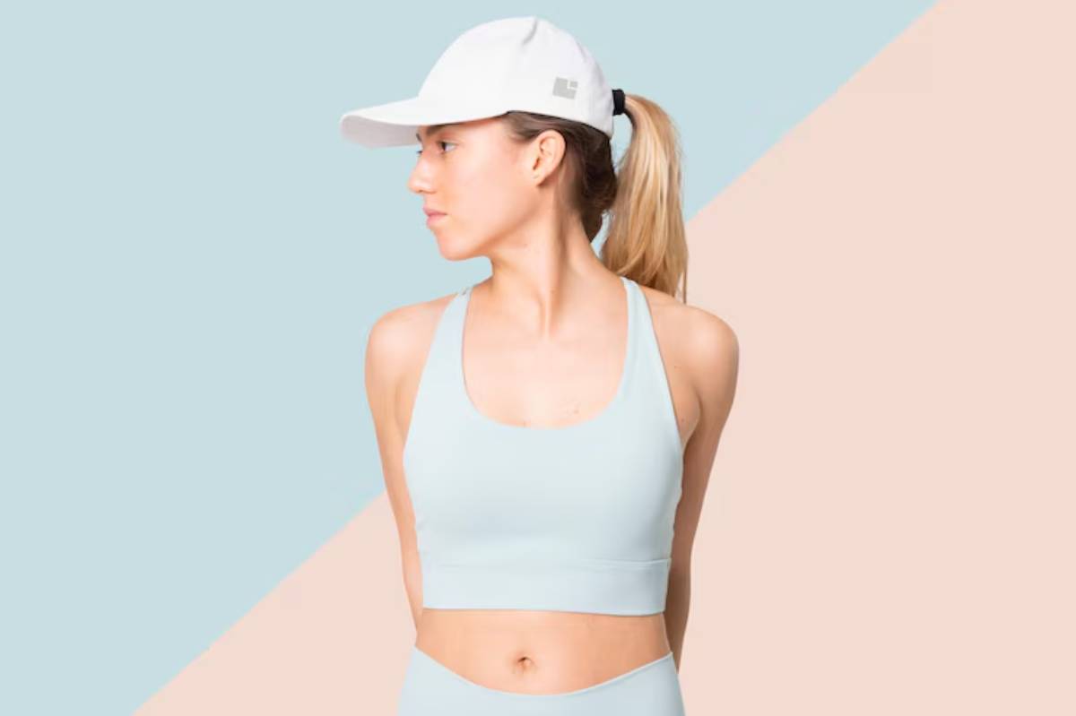 A woman in a light blue sports bra and cap poses against a pastel blue and pink background, showcasing an athletic vibe.