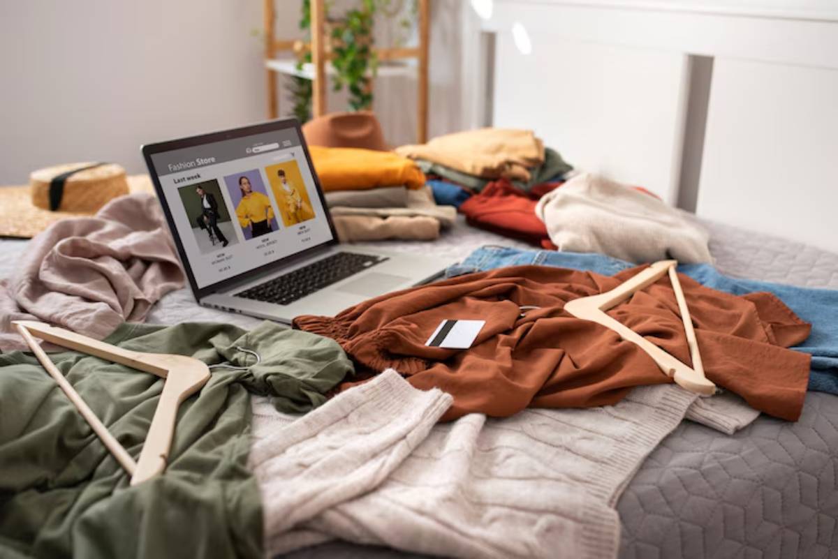 A laptop displays a fashion store website amidst a messy bed covered with colorful clothing and wooden hangers.