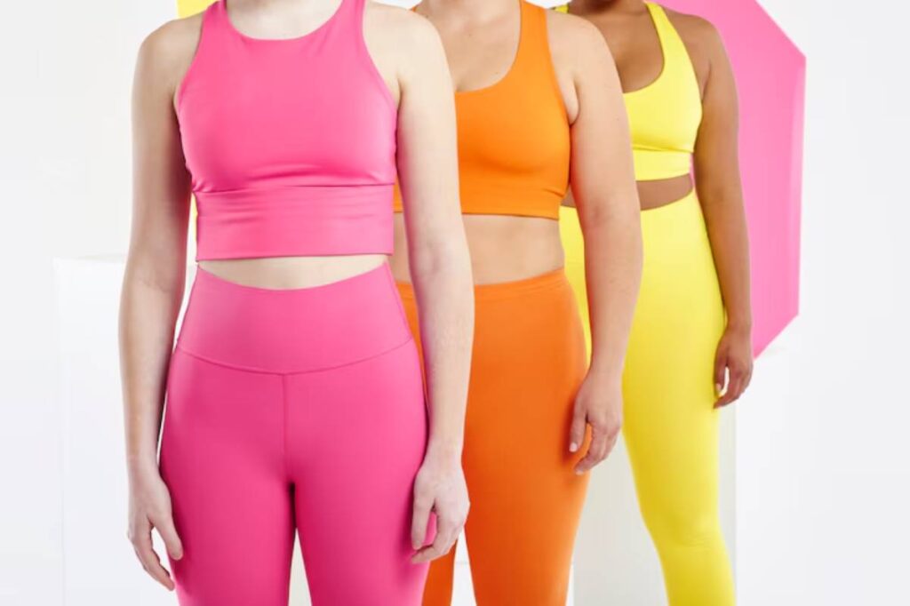 Three women in vibrant athletic wear: pink, orange, and yellow, stand side by side against a colorful backdrop, showcasing confidence and style.