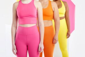 Three women in vibrant athletic wear: pink, orange, and yellow, stand side by side against a colorful backdrop, showcasing confidence and style.