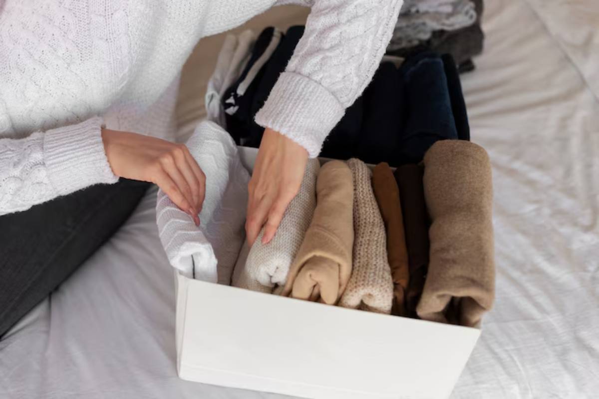 A person wearing a white sweater neatly folds and organizes soft, stacked clothes in a white storage box on a bed.