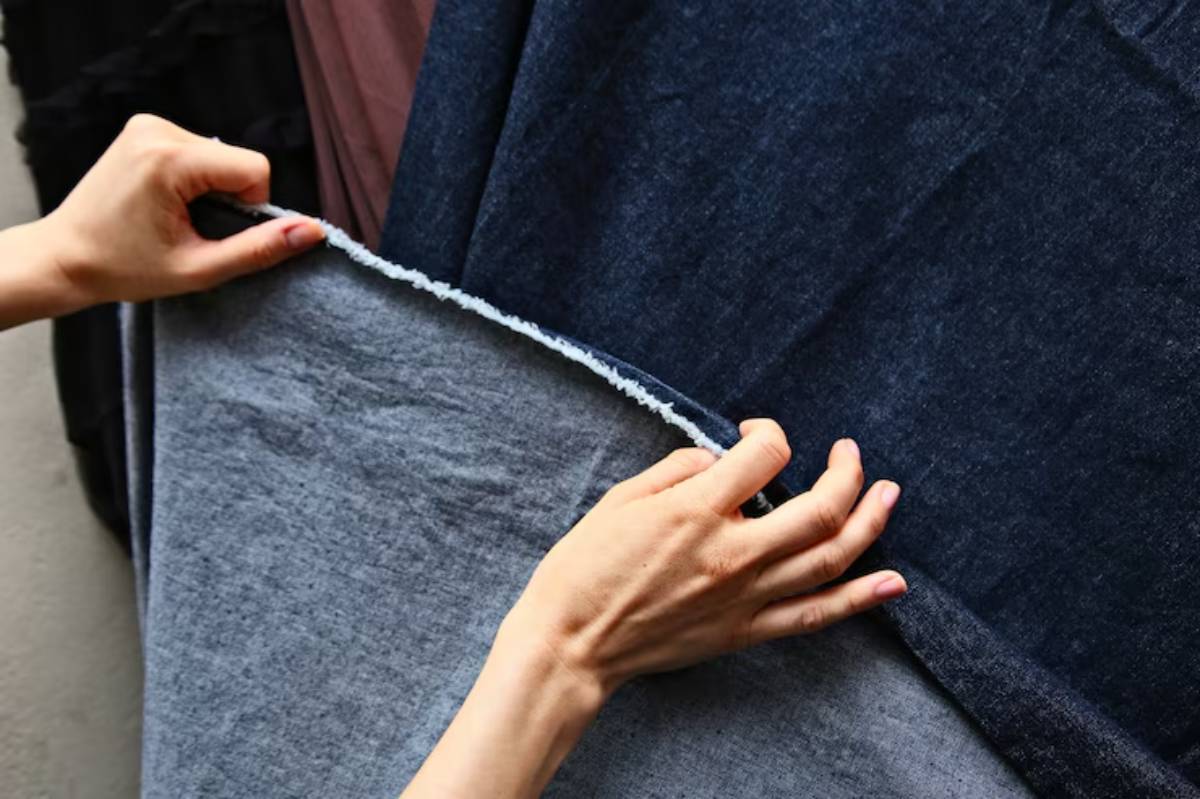 A hand holds a piece of fabric, revealing a dark blue textile layered over a lighter gray one, showcasing different fabric textures.