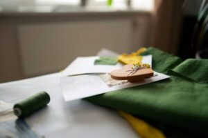 Close-up of sewing materials including green fabric, colored thread, sketches, and a pin cushion with pins on a work table.