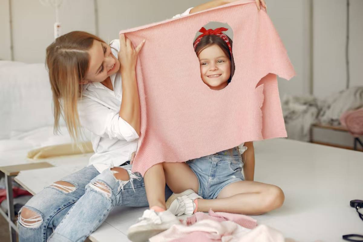 Two girls playfully hold a pink sweater, sitting on a table surrounded by soft fabric, in a cozy indoor setting.