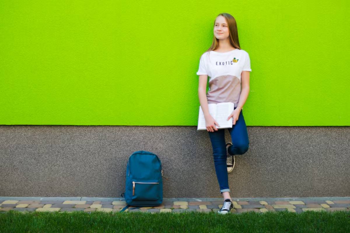 A young person in a casual outfit stands against a vibrant green wall, holding a notebook, with a blue backpack beside them.