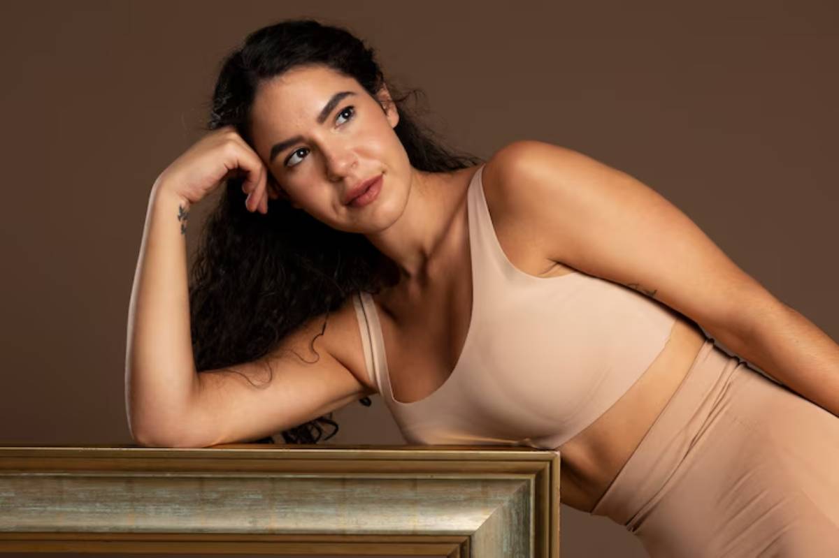 A woman with long, curly hair rests her chin on her hand, wearing a beige crop top, leaning against a gold-framed rectangular surface.