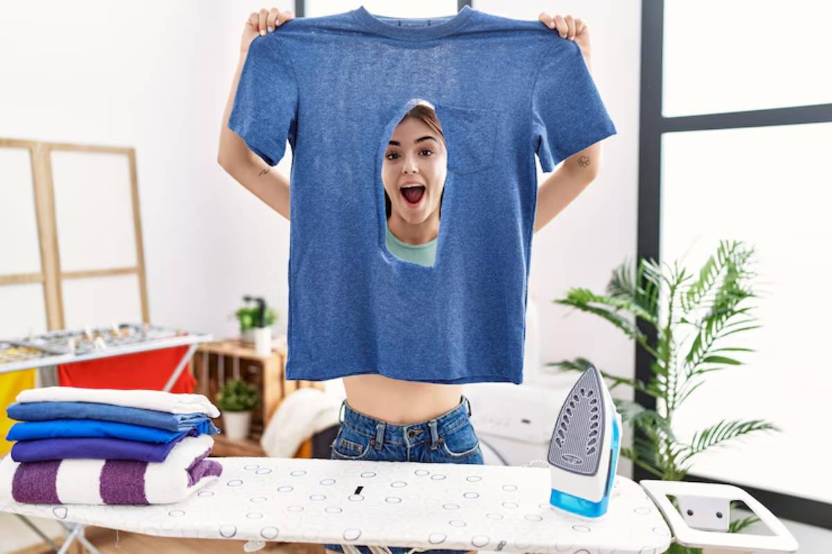 A person holding up a blue t-shirt in front of an ironing board with neatly stacked towels and a blue iron nearby.