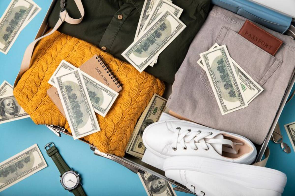 A suitcase packed with a yellow sweater, green shirt, gray pants, white sneakers, a notebook, and scattered dollar bills.