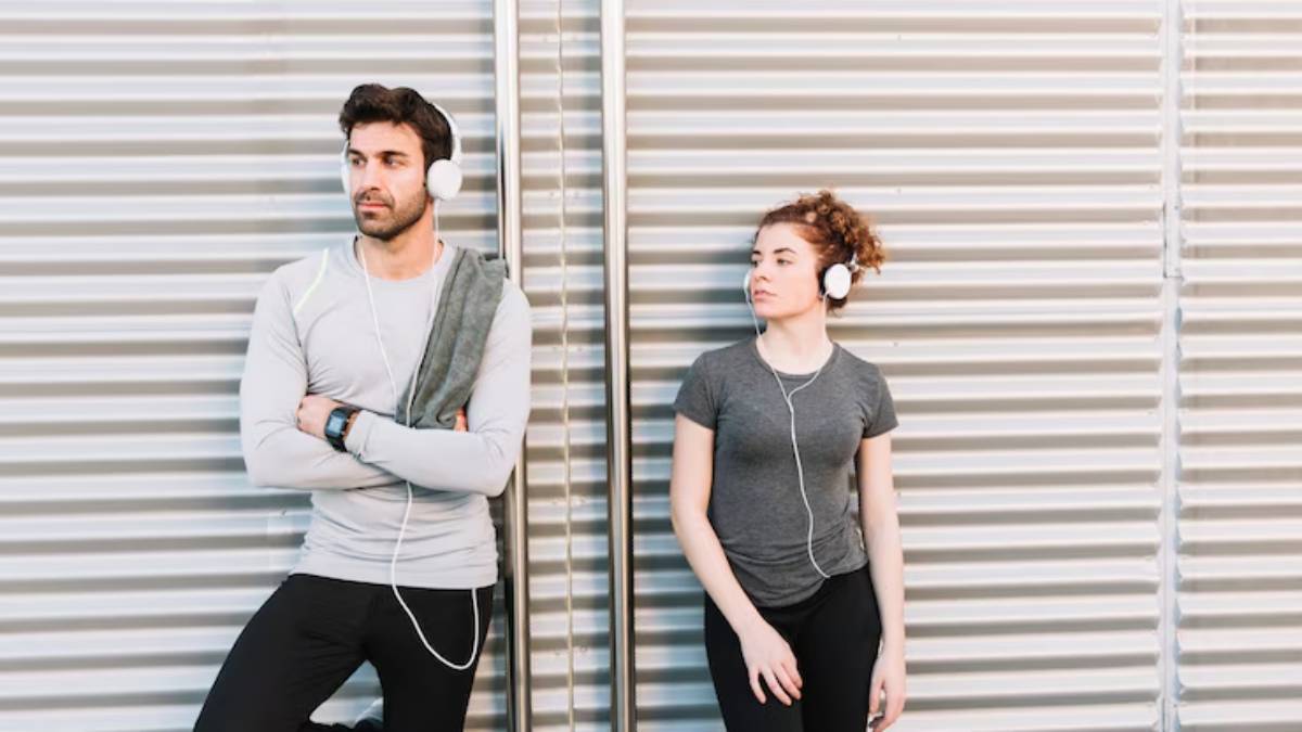 A man and a woman stand against a corrugated metal wall, both wearing headphones and athletic clothing, lost in their music.