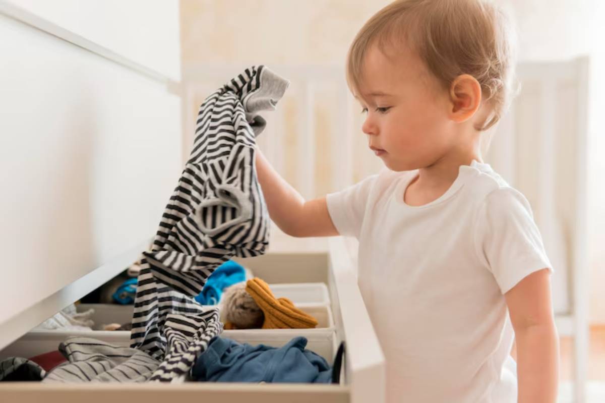 A child in a white shirt pulls out a striped shirt from a drawer filled with colorful clothes and toys. Bright indoor setting.
