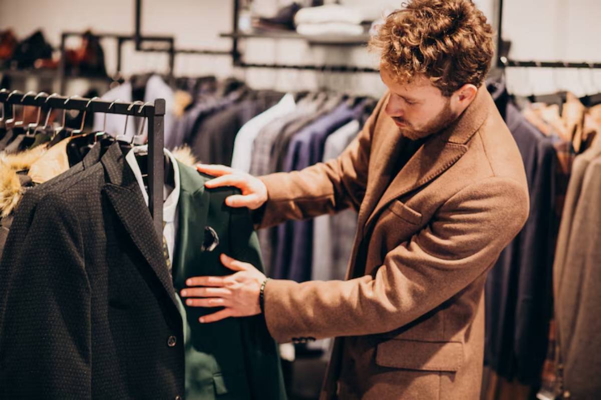 A man in a stylish brown coat examines a green blazer on a clothing rack in a boutique filled with various suits.