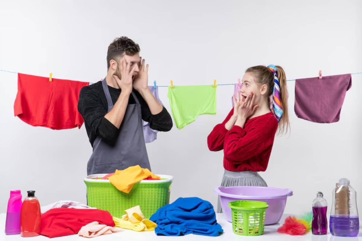 A man and a woman stand in front of colorful laundry, with clothing hanging on a line and laundry supplies on a table between them.