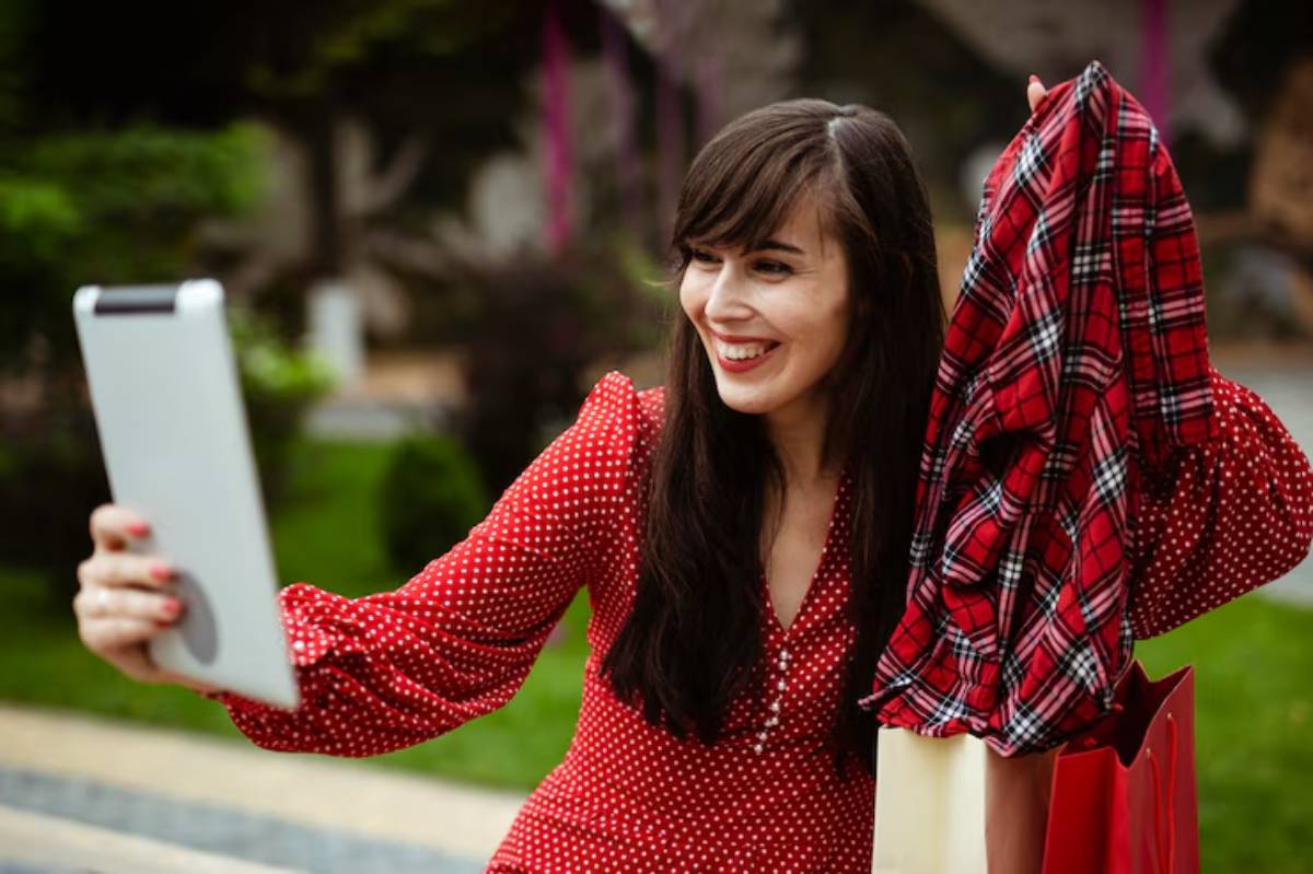 A woman in a red polka dot dress takes a selfie while holding a red plaid shirt and a shopping bag in a lush outdoor setting.