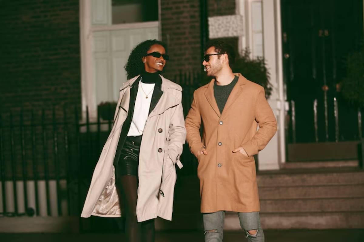 A couple walks together in stylish winter outfits; one in a beige trench coat and the other in a tan overcoat against a brownstone backdrop.