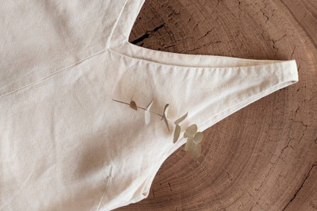 A light beige fabric piece with a v-shaped neckline, adorned with a sprig of eucalyptus against a textured wood background.