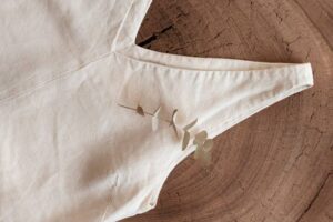 A light beige fabric piece with a v-shaped neckline, adorned with a sprig of eucalyptus against a textured wood background.