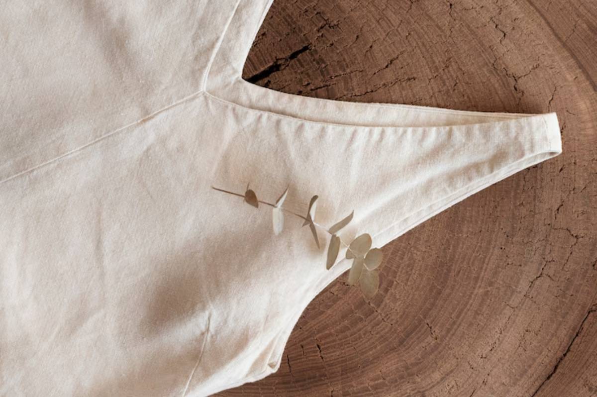 A light beige fabric piece with a v-shaped neckline, adorned with a sprig of eucalyptus against a textured wood background.