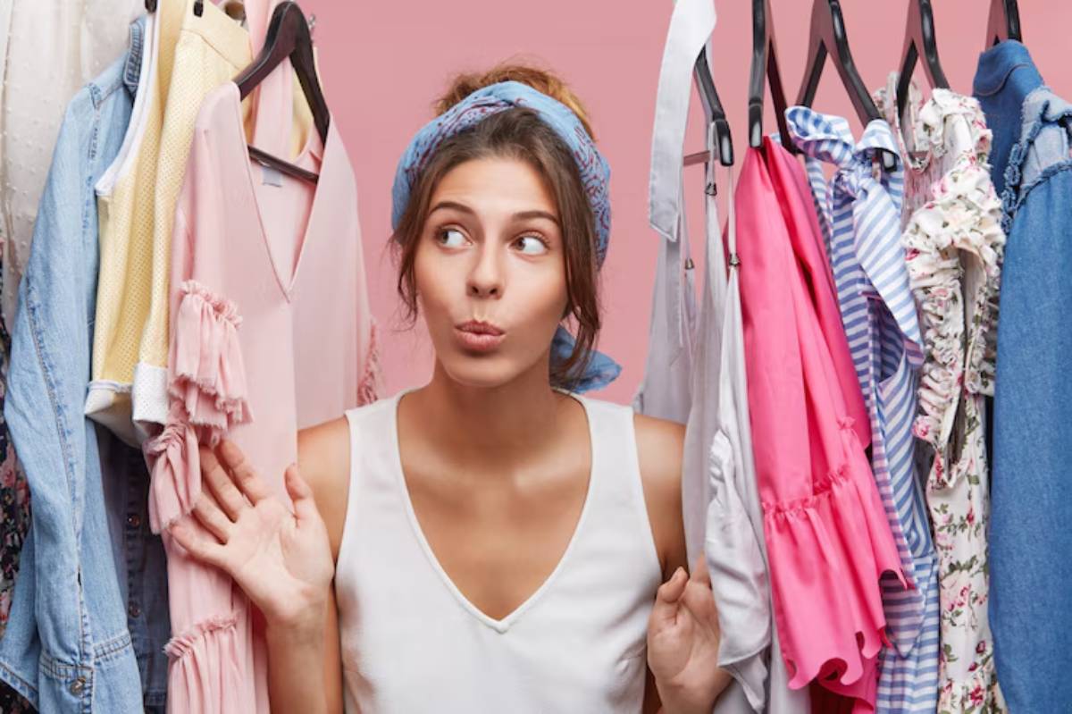 A woman in a tank top stands amid colorful clothing on hangers against a pink backdrop, looking towards the camera with a friendly gesture.