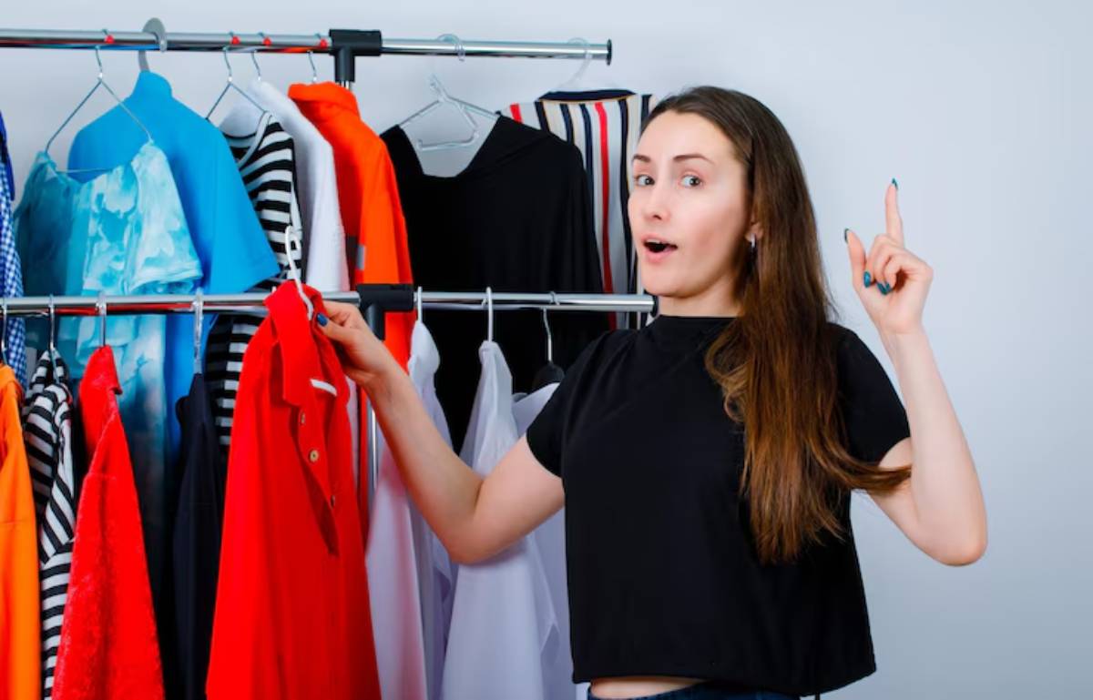 A woman in a black shirt gestures while holding a red shirt, surrounded by a colorful array of clothing on hangers.