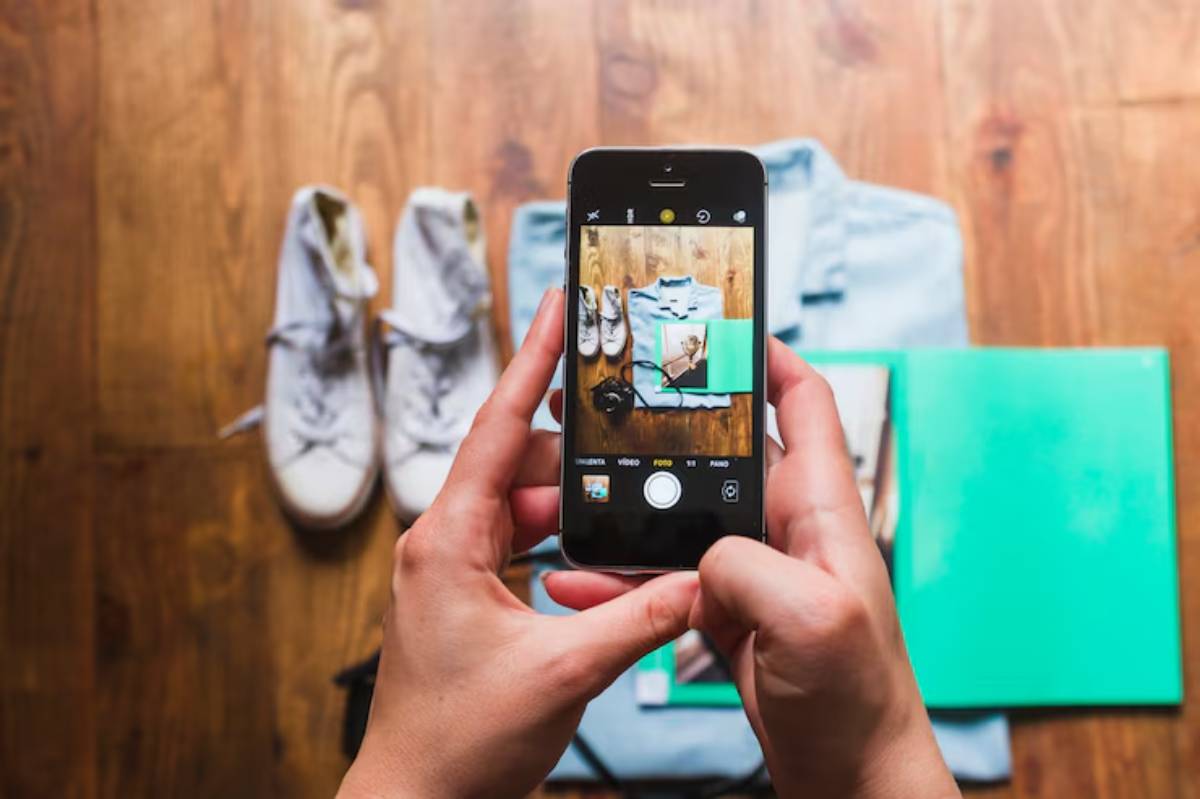 A person holding a smartphone takes a flat lay photo of clothing, shoes, and accessories on a wooden floor.