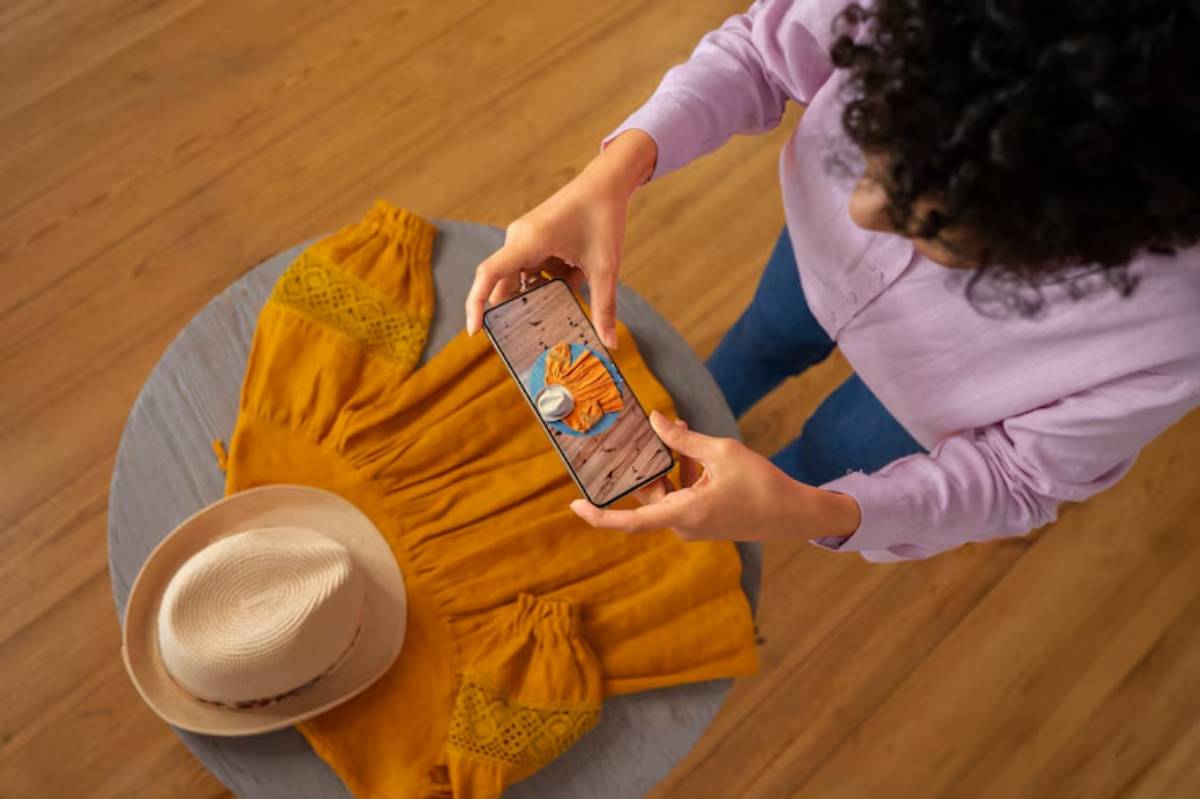A person holds a smartphone above a mustard yellow dress and straw hat, likely capturing a photo in a wooden-floored setting.